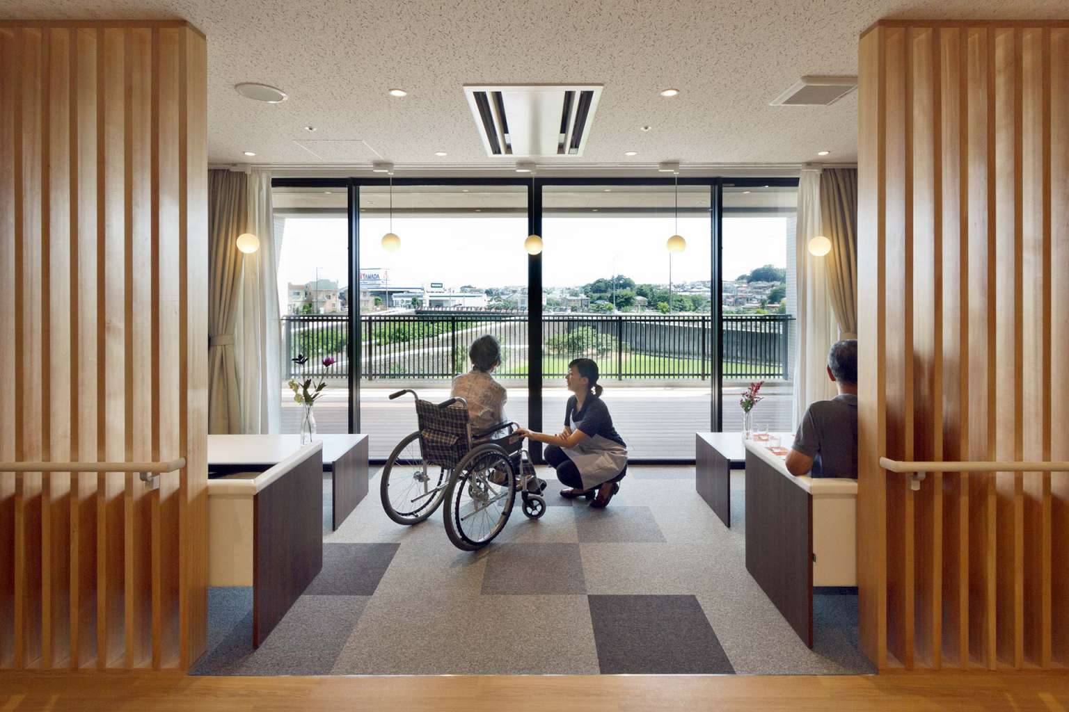 車椅子利用者を含む入居者がテラスを眺める共用スペース。木製装飾と大型窓が特徴