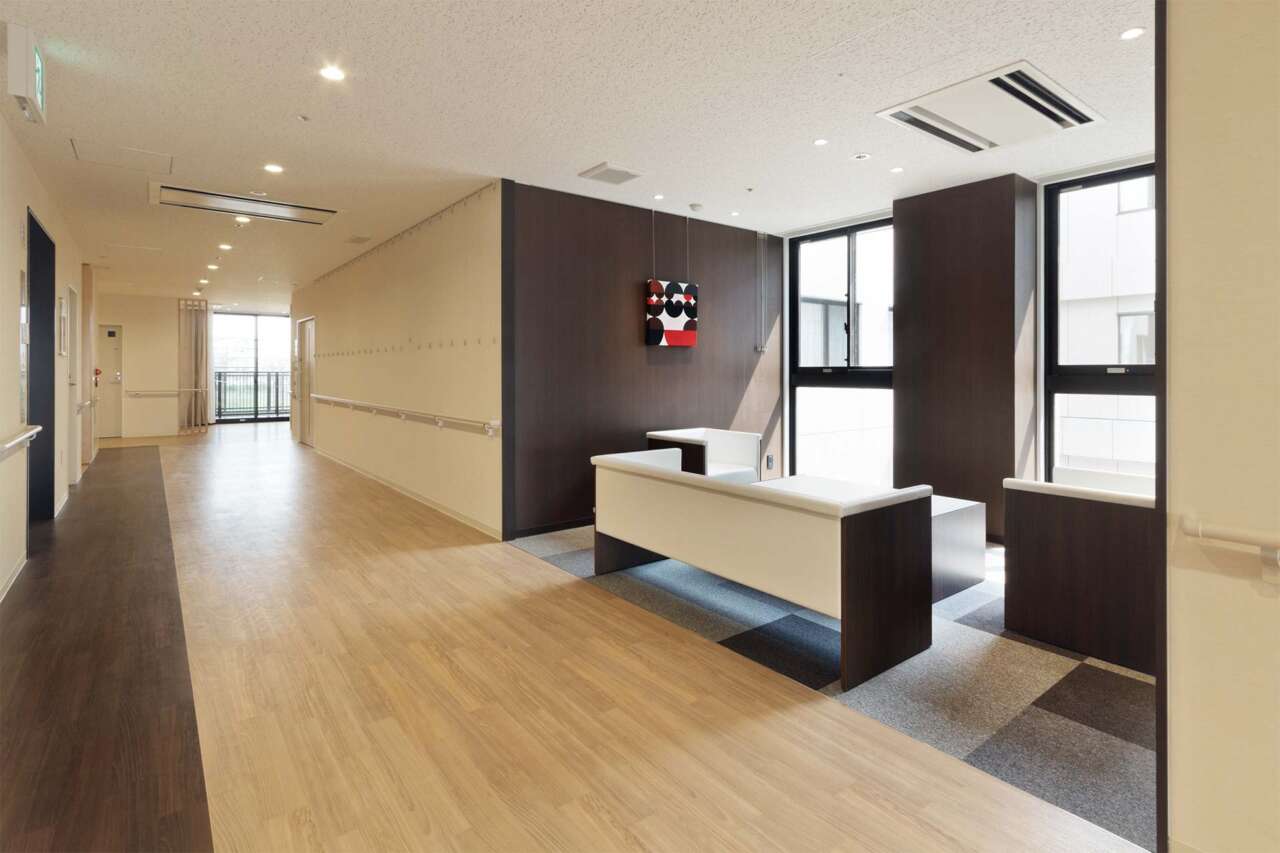 モダンな受付カウンターと廊下を備えた明るいロビー空間。木調床と落ち着いた色調の壁が特徴。